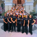 El equipo de capacitación de la Empresa Zulueta posa en primer plano, todos vestidos con camisetas negras con el logotipo de la firma. Detrás de ellos, en una elegante escalera, se encuentran las 17 embajadoras de la Feria Tabasco, vestidas con trajes regionales de blusa bordada con flores y falda.