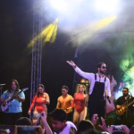 En El Escenario, Chico Che Chico Aparece En El Centro Con Su Característico Overol Negro Y Camisa Blanca, Con Un Brazo Extendido Hacia El Público. Lo Acompañan Músicos Con Guitarras Y Bailarinas Con Vestuarios Rojos. Luces Amarillas De Escenario Iluminan La Escena Bajo Una Estructura De Metal. Al Frente Se Observan Las Manos Del Público Sosteniendo Teléfonos Celulares.