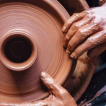 Una vista aérea picada (cenital) captura un primer plano extremo de las manos de un artesano alfarero cubiertas de barro arcilloso de color terracota.