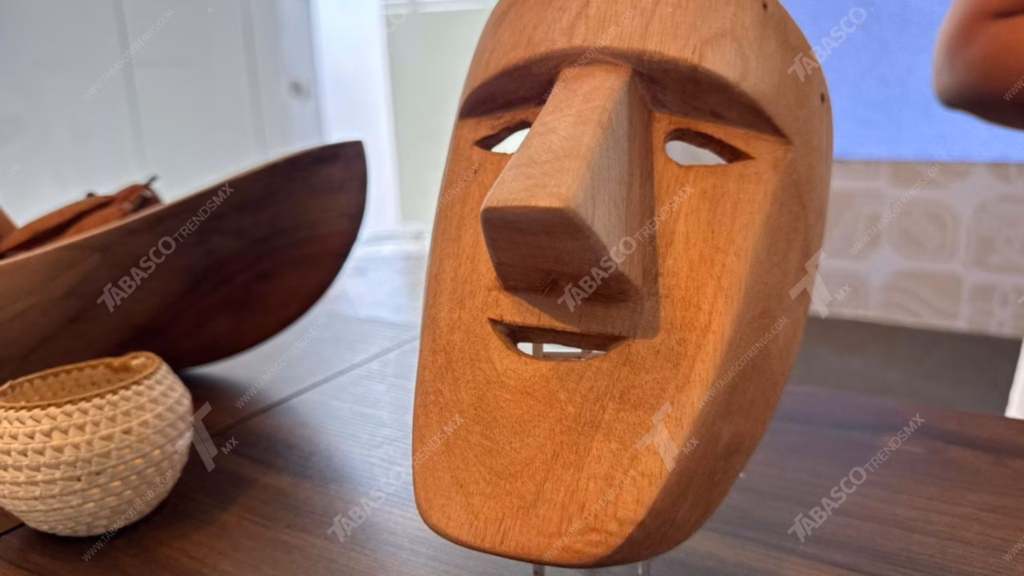 Ganadores Del Reconocimiento De Excelencia Artesanal 2026 En Tabasco 71 Primer Plano De Una Máscara Artesanal Tallada En Una Sola Pieza De Madera De Color Claro, Con Una Expresión Neutra Y Aberturas Para Los Ojos Y La Boca, Mostrada En Un Ángulo De Tres Cuartos. La Máscara Está Sobre Una Mesa De Madera. En El Fondo, Desenfocado A La Izquierda, Hay Un Cuenco De Madera Más Grande Y Una Pequeña Cesta Tejida De Color Claro, Lo Que Sugiere Una Exhibición De Artesanías Regionales.