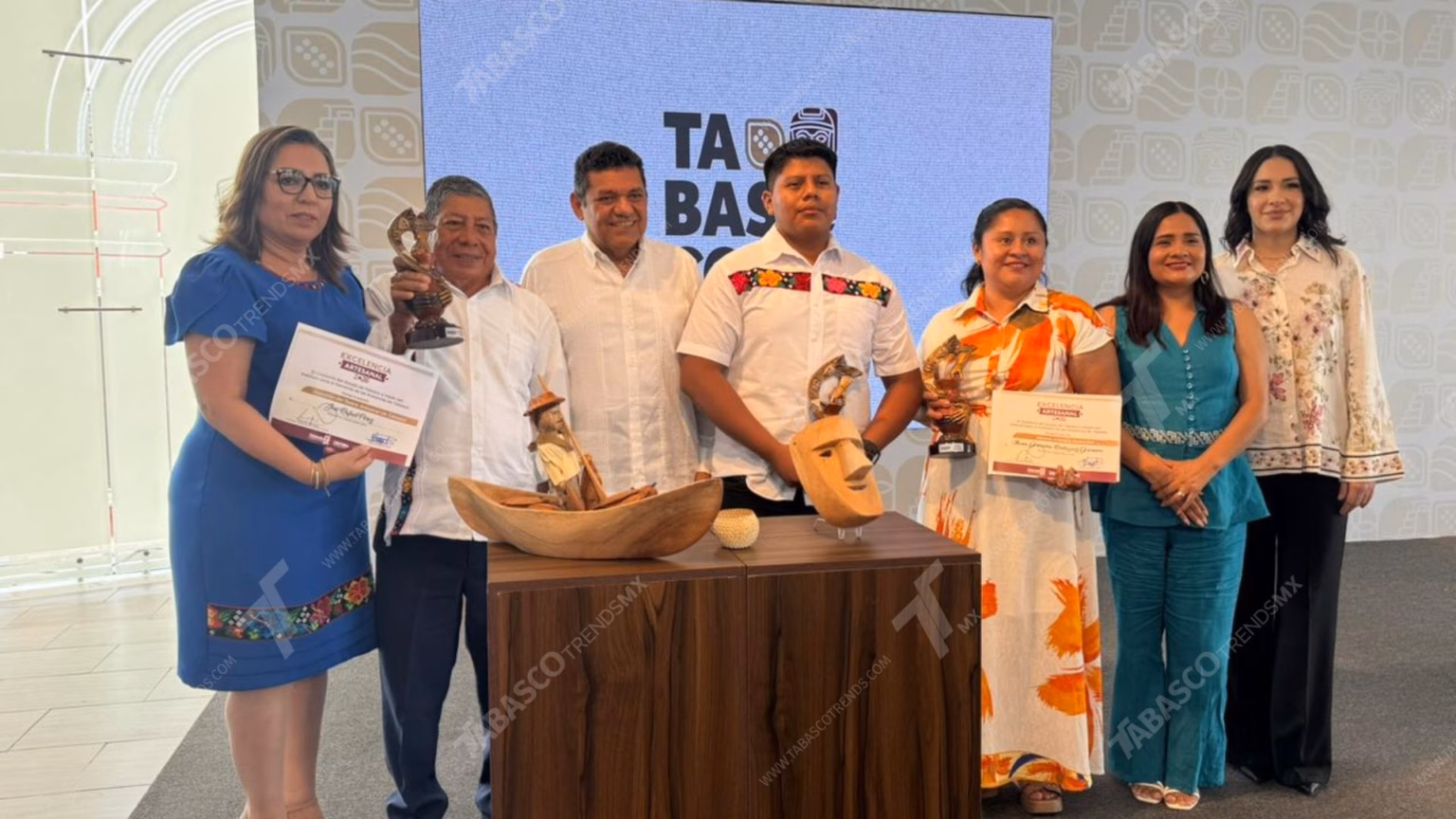 Ganadores Del Reconocimiento De Excelencia Artesanal 2026 En Tabasco 69 Fotografía Grupal Durante La Presentación Del Reconocimiento De Excelencia Artesanal 2026 En Tabasco. En El Centro, Los Artesanos Ganadores Sostienen Sus Reconocimientos Y Trofeos Junto A Autoridades Estatales. Al Frente, Sobre Una Mesa De Madera, Se Exhiben Piezas Artesanales: Una Canoa Tallada Con Un Pescador Y Una Máscara Tradicional De La Danza Del Pochó. El Fondo Muestra El Logotipo De &Quot;Tabasco&Quot; Y Un Patrón De Iconografía Regional.