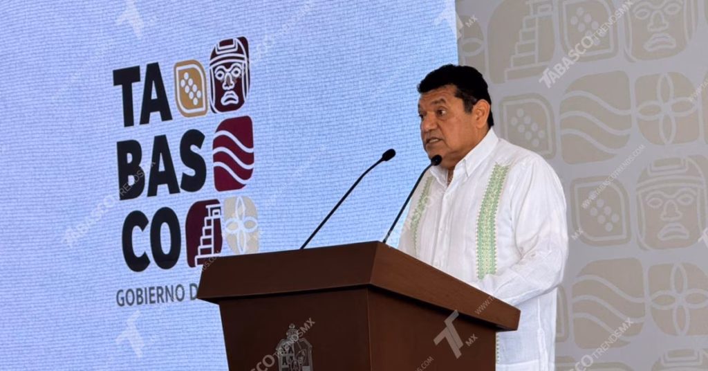 El Gobernador de Tabasco, Javier May Rodríguez, de pie tras un podio de madera durante una conferencia de prensa. Viste una guayabera blanca de manga larga con detalles de bordado verde en el pecho.