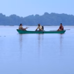 aisaje panorámico de Potonchán en los Pantanos de Centla, Tabasco. Se observa una canoa verde con tres personas navegando sobre un espejo de agua sereno bajo un cielo nublado. Al fondo, una densa línea de manglares y selva completa el horizonte, transmitiendo una atmósfera de paz y conexión con la naturaleza.