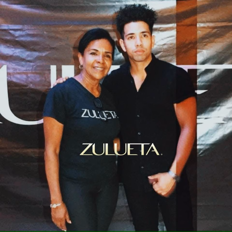Fotografía de Rossana Zulueta y Félix Zulueta posando juntos frente a un fondo oscuro con el logotipo "ZULUETA". Rossana viste una camiseta negra con el nombre de la empresa y Félix una camisa negra de botones. Ambos representan el liderazgo de la firma de capacitación.
