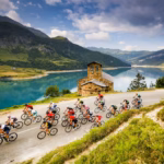 Una toma aérea de un pelotón de ciclistas de élite compitiendo en una carretera sinuosa de montaña durante una etapa de L'Étape by Tour de France. El grupo de ciclistas, con uniformes multicolores, avanza sobre el asfalto frente a un paisaje espectacular.