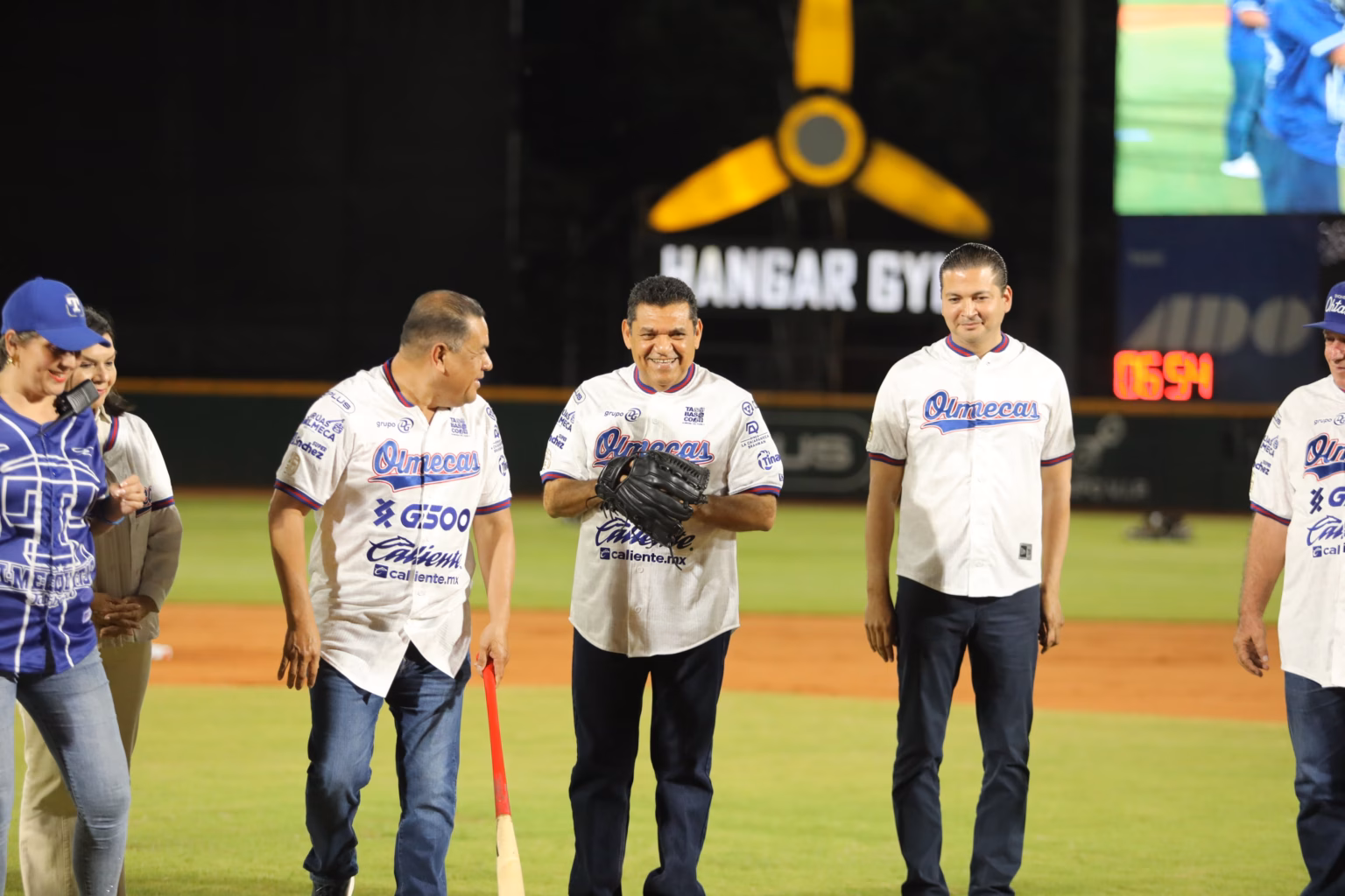 Una Fotografía En Plano Medio Tomada De Noche En El Estadio Centenario 27 De Febrero. El Gobernador Javier May Rodríguez Está En El Centro Del Diamante De Béisbol, Sonriendo Y Usando Un Guante De Béisbol Negro En La Mano Izquierda.