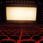 Una Fotografía En Gran Angular Que Muestra El Interior De Una Sala De Cine Vacía Con Una Atmósfera Cinematográfica Y Solemne. La Perspectiva Se Sitúa Desde Las Filas Traseras, Mirando Hacia Una Pantalla Blanca De Gran Formato Que Ilumina El Espacio.