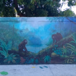 Un Mural Artístico Pintado Sobre Una Barda Exterior Que Representa Una Selva Tropical. En El Centro Destacan Dos Monos Saraguatos Sobre Ramas De Árboles, Rodeados De Vegetación Verde Intensa, Helechos Y Flores Exóticas. Al Fondo Se Insinúa Un Cuerpo De Agua Bajo Una Luz Azulada.