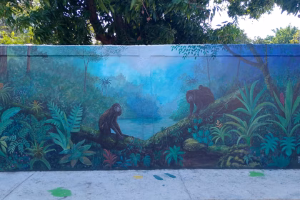 Un Mural Artístico Pintado Sobre Una Barda Exterior Que Representa Una Selva Tropical. En El Centro Destacan Dos Monos Saraguatos Sobre Ramas De Árboles, Rodeados De Vegetación Verde Intensa, Helechos Y Flores Exóticas. Al Fondo Se Insinúa Un Cuerpo De Agua Bajo Una Luz Azulada.