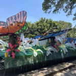Fotografía A Plena Luz Del Día De Un Carro Alegórico Monumental Del Municipio De Cunduacán Estacionado En Una Avenida. El Diseño Destaca Por Grandes Figuras De Mariposas Con Alas Translúcidas En Tonos Naranja, Azul Y Verde, Que Parecen Sobrevolar Un Jardín De Flores Blancas Gigantes De Tela O Papel.