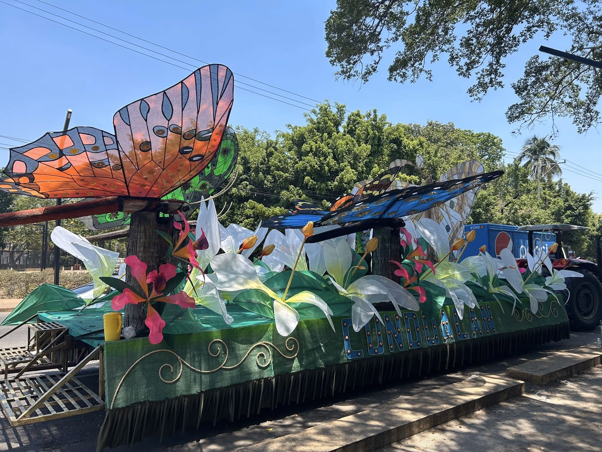 Fotografía A Plena Luz Del Día De Un Carro Alegórico Monumental Del Municipio De Cunduacán Estacionado En Una Avenida. El Diseño Destaca Por Grandes Figuras De Mariposas Con Alas Translúcidas En Tonos Naranja, Azul Y Verde, Que Parecen Sobrevolar Un Jardín De Flores Blancas Gigantes De Tela O Papel.