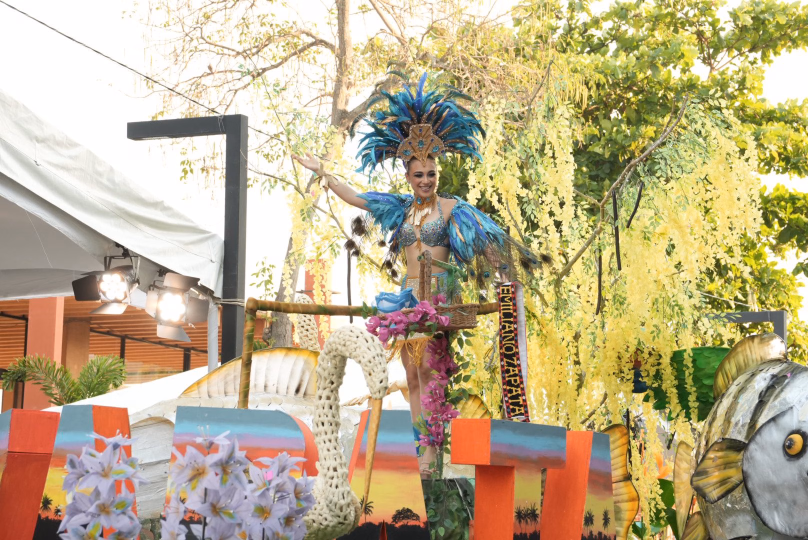 Mariú Mendoza Cabrera, Embajadora De Emiliano Zapata, Viste Un Traje De Gala Con Plumas Azul Turquesa Y Pedrería, Saludando Desde Un Carro Alegórico Decorado Con Flores De Lluvia De Oro Y Elementos Que Representan La Fauna Del Río Usumacinta.
