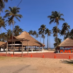 Fotografía Del Acceso Principal A Playa Del Sol En Paraíso, Tabasco, Bajo Un Cielo Azul Despejado. En Primer Plano Hay Un Camino De Arena Y Una Franja Pavimentada De Color Rojo Delineada Con Conos De Tráfico Naranjas Para Guiar El Flujo De Visitantes.