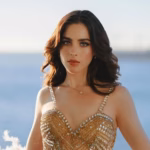 Retrato De Fátima Bosch, Miss Universo 2025, Luciendo Un Vestido De Gala Dorado Con Pedrería Y Plumas Blancas, En Un Entorno Costero Con El Mar Y Un Barco Al Fondo, Capturando Un Momento De Serenidad Y Sofisticación.