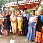 Fotografía Grupal De Cocineros Tradicionales Con Delantales Y Trajes Típicos Tabasqueños, Junto A Autoridades Estatales, Posando Frente A Un Puesto De Comida Tradicional Con Techo De Teja. Se Observa Un Ambiente Festivo Y Cultural Al Aire Libre.