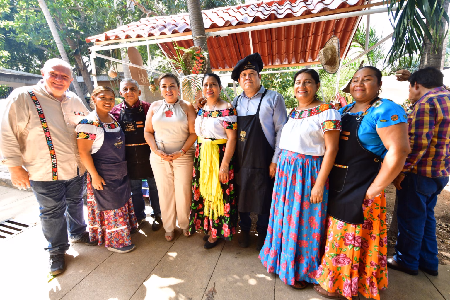Fotografía Grupal De Cocineros Tradicionales Con Delantales Y Trajes Típicos Tabasqueños, Junto A Autoridades Estatales, Posando Frente A Un Puesto De Comida Tradicional Con Techo De Teja. Se Observa Un Ambiente Festivo Y Cultural Al Aire Libre.