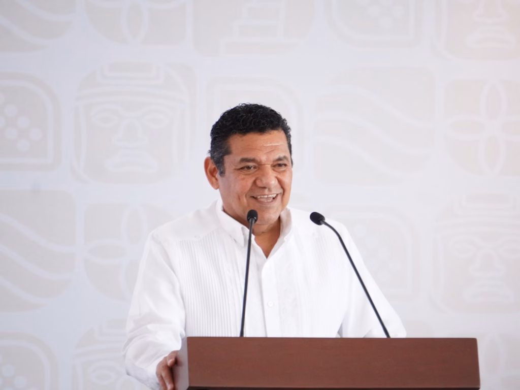 Javier May Reporta Saldo Blanco Y Baja De Homicidios En Tabasco 2026 71 Retrato De Medio Cuerpo Del Gobernador Javier May Rodríguez, Un Hombre De Mediana Edad Con Cabello Oscuro Y Rizado, Vistiendo Una Guayabera Blanca Tradicional. Se Encuentra De Pie Detrás De Un Podio De Madera Marrón Con Dos Micrófonos, Sonriendo Durante Un Discurso. El Fondo Es De Color Claro Con Patrones Gráficos Tenues De Iconografía Prehispánica De Tabasco.