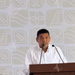 El Gobernador De Tabasco, Javier May Rodríguez, De Pie Tras Un Podio Con Micrófonos, Vistiendo Una Guayabera Blanca Tradicional.
