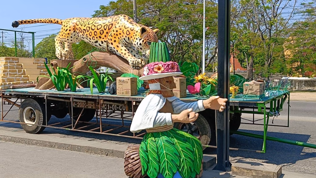 Un Plano Medio-Bajo A Nivel De Calle Muestra El Carro Alegórico En Proceso Del Municipio De Tenosique Para La Feria Tabasco 2026. La Plataforma Del Camión Está Decorada Con Vibrantes Elementos Selváticos En Cartonería.
