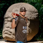 El Beisbolista Posa Frente A Una Monumental Cabeza Colosal Olmeca. Viste La Nueva Camisola De Los Olmecas De Tabasco Denominada &Quot;El Origen De Todo&Quot;, En Un Tono Café Tierra Con Detalles En Color Crema Y Logotipos De Patrocinadores. Sostiene Un Bate De Madera Roja Sobre Sus Hombros Y Usa Una Gorra Café Con El Logotipo De La &Quot;T&Quot; Entrelazada. El Fondo Es De Vegetación Selvática Verde, Creando Un Contraste Cultural Y Deportivo.