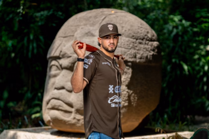 El Beisbolista Posa Frente A Una Monumental Cabeza Colosal Olmeca. Viste La Nueva Camisola De Los Olmecas De Tabasco Denominada &Quot;El Origen De Todo&Quot;, En Un Tono Café Tierra Con Detalles En Color Crema Y Logotipos De Patrocinadores. Sostiene Un Bate De Madera Roja Sobre Sus Hombros Y Usa Una Gorra Café Con El Logotipo De La &Quot;T&Quot; Entrelazada. El Fondo Es De Vegetación Selvática Verde, Creando Un Contraste Cultural Y Deportivo.