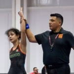 Fotografía De Una Competencia De Lucha Libre Olímpica En Un Gimnasio Techado. En El Centro, Un Árbitro Con Uniforme Blanco Levanta El Brazo De Una Joven Luchadora Que Viste El Uniforme De La Selección De México (Color Negro Con Detalles En Verde Y Rojo) En Señal De Victoria. A Su Lado Se Encuentra Su Oponente Con El Uniforme De Argentina (Azul Y Blanco). Ambas Están Sobre Un Colchón De Lucha Circular Azul Y Naranja Bajo Luces Profesionales De Pabellón Deportivo.