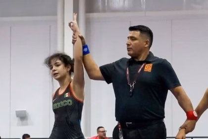 Fotografía De Una Competencia De Lucha Libre Olímpica En Un Gimnasio Techado. En El Centro, Un Árbitro Con Uniforme Blanco Levanta El Brazo De Una Joven Luchadora Que Viste El Uniforme De La Selección De México (Color Negro Con Detalles En Verde Y Rojo) En Señal De Victoria. A Su Lado Se Encuentra Su Oponente Con El Uniforme De Argentina (Azul Y Blanco). Ambas Están Sobre Un Colchón De Lucha Circular Azul Y Naranja Bajo Luces Profesionales De Pabellón Deportivo.