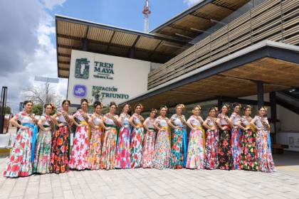 Las 17 Embajadoras De La Feria Tabasco 2026 Posan Alineadas Frente A La Moderna Fachada De La Estación El Triunfo Del Tren Maya. Todas Visten El Traje Regional De Gala Tabasqueño, Que Consiste En Blusas Blancas Con Tiras Bordadas Y Faldas Largas Floreadas De Distintos Colores. Llevan Puestas Sus Bandas Municipales Y Flores En El Cabello. El Edificio De La Estación Tiene Un Diseño Contemporáneo Con Estructuras De Madera Y El Logotipo Oficial Del Tren Maya.