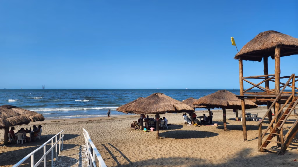 ¡Enamórate De Tabasco! Siete Playas Certificadas Te Esperan Esta Semana Santa 2026 70 Vista Panorámica De Playa Del Sol En Paraíso, Tabasco, Durante Un Día Soleado Con Cielo Despejado. En La Arena Clara Se Distribuyen Varias Palapas De Techo De Paja Que Ofrecen Sombra A Grupos De Visitantes Sentados En Mesas Y Sillas. A La Derecha, Destaca Una Torre De Vigilancia De Madera De Dos Niveles Con Una Bandera Amarilla.