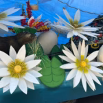 Un Primer Plano De Los Detalles Decorativos De Un Carro Alegórico. En La Escena Destacan Flores De Loto Blancas De Gran Tamaño Con Centros Amarillos, Hojas Verdes Circulares Y Una Figura De Un Manatí Marrón De Textura Realista. Al Fondo Se Observa Una Tortuga Decorada Con Patrones Coloridos Y Pequeñas Flores Rojas, Junto A Un Huevo Del Que Emerge Una Pequeña Tortuga Amarilla. Todo El Conjunto Está Bajo Una Carpa Protectora Azul, Mostrando El Trabajo Artesanal De Jonuta Para La Feria Tabasco 2026.