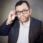 Retrato En Primer Plano Del Actor Mexicano Silverio Palacios. Aparece Con Una Expresión Seria Y Analítica, Ajustándose Sus Lentes De Armazón Negro.