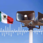 Primer Plano De Dos Altavoces De Alerta Sísmica Color Gris Montados Sobre Un Poste Blanco, Con La Bandera De México Ondeando Al Fondo Bajo Un Cielo Azul Despejado. Una Representación Gráfica De Una Onda Sonora Blanca Cruza La Imagen, Simbolizando La Activación De La Alarma Para El Simulacro Nacional 2026.