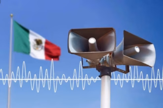 Primer Plano De Dos Altavoces De Alerta Sísmica Color Gris Montados Sobre Un Poste Blanco, Con La Bandera De México Ondeando Al Fondo Bajo Un Cielo Azul Despejado. Una Representación Gráfica De Una Onda Sonora Blanca Cruza La Imagen, Simbolizando La Activación De La Alarma Para El Simulacro Nacional 2026.