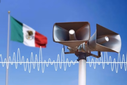 Primer Plano De Dos Altavoces De Alerta Sísmica Color Gris Montados Sobre Un Poste Blanco, Con La Bandera De México Ondeando Al Fondo Bajo Un Cielo Azul Despejado. Una Representación Gráfica De Una Onda Sonora Blanca Cruza La Imagen, Simbolizando La Activación De La Alarma Para El Simulacro Nacional 2026.