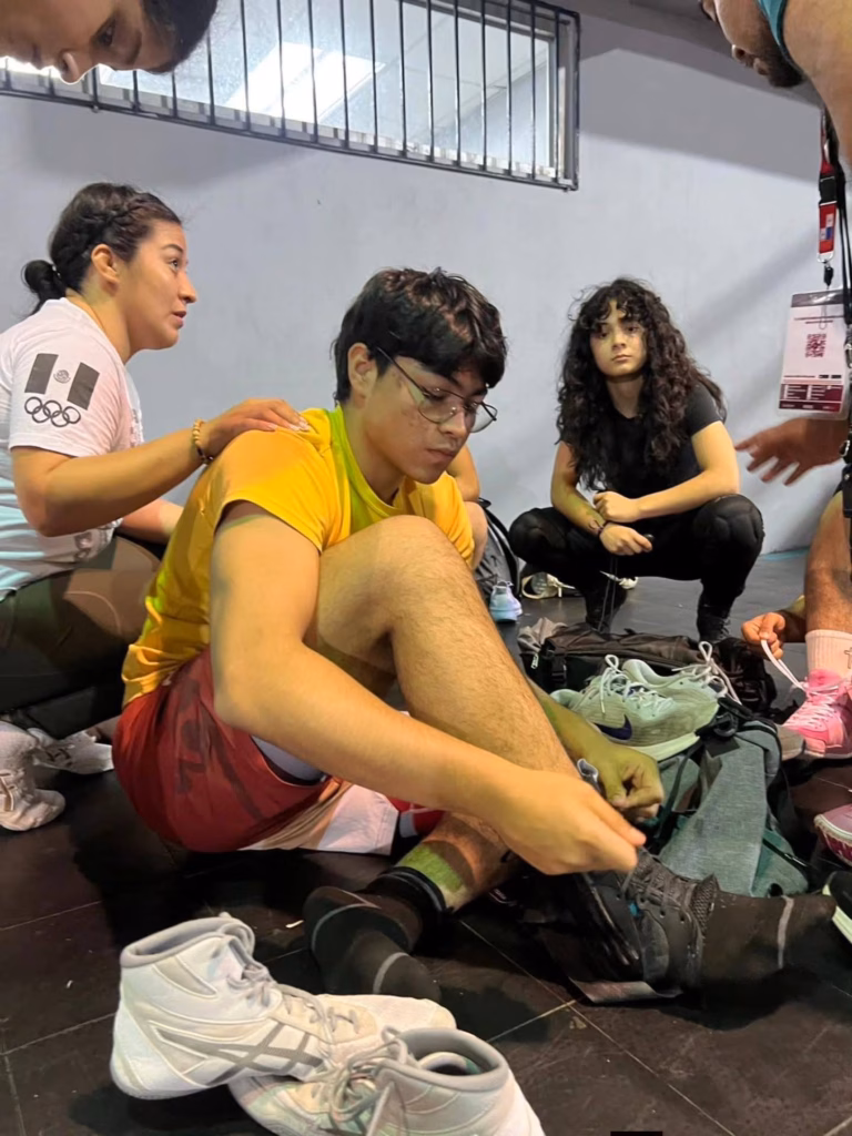 ¡Orgullo Tabasqueño En Panamá! Valentina Padrón Conquista El Bronce En El Panamericano De Luchas Asociadas U17 71 Toma De Ángulo Bajo Que Muestra A La Atleta Valentina Padrón Sentada En El Suelo De Un Gimnasio, Concentrada Mientras Ajusta Las Agujetas De Sus Botas De Lucha. Viste Una Playera Deportiva Amarilla. Al Fondo Se Observa A Su Entrenadora Brindándole Apoyo Y Otros Atletas En El Área De Calentamiento.