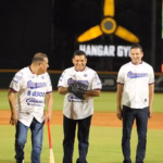 Una Fotografía En Plano Medio Tomada De Noche En El Estadio Centenario 27 De Febrero. El Gobernador Javier May Rodríguez Está En El Centro Del Diamante De Béisbol, Sonriendo Y Usando Un Guante De Béisbol Negro En La Mano Izquierda.