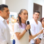 Fotografía En Plano Medio Y Toma De Tres Cuartos En Interiores Iluminados. En El Centro, La Presidenta Claudia Sheinbaum Pardo Habla Sosteniendo Un Micrófono Negro Con La Mano Derecha Y Gesticula Con La Izquierda; Viste Un Vestido Blanco Con Detalles Bordados En Color Morado En El Cuello. A Su Derecha, En Primer Plano, Un Hombre Con Guayabera Blanca De Manga Larga Escucha Con Atención Mirando Hacia El Frente. En El Fondo, De Forma Ligeramente Difuminada, Se Aprecian Otras Personas Alineadas, Incluyendo A Un Hombre Joven Con Guayabera Blanca Y A Una Mujer Con Lentes Y Blusa Blanca.