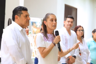 Fotografía En Plano Medio Y Toma De Tres Cuartos En Interiores Iluminados. En El Centro, La Presidenta Claudia Sheinbaum Pardo Habla Sosteniendo Un Micrófono Negro Con La Mano Derecha Y Gesticula Con La Izquierda; Viste Un Vestido Blanco Con Detalles Bordados En Color Morado En El Cuello. A Su Derecha, En Primer Plano, Un Hombre Con Guayabera Blanca De Manga Larga Escucha Con Atención Mirando Hacia El Frente. En El Fondo, De Forma Ligeramente Difuminada, Se Aprecian Otras Personas Alineadas, Incluyendo A Un Hombre Joven Con Guayabera Blanca Y A Una Mujer Con Lentes Y Blusa Blanca.
