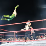 Fotografía De Acción Durante Una Función De Lucha Libre En El Foro Tabasco De La Feria Tabasco 2026. Un Luchador Con Equipo Verde Y Amarillo Realiza Un Espectacular Vuelo Desde La Tercera Cuerda Hacia Su Oponente, Quien Lo Espera De Pie Sobre El Ring. El Referee Y El Público, Que Llena Las Gradas Bajo La Noche, Observan La Maniobra. Las Cuerdas Del Ring Son Rojas Y La Iluminación Destaca El Movimiento Aéreo.