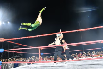Fotografía De Acción Durante Una Función De Lucha Libre En El Foro Tabasco De La Feria Tabasco 2026. Un Luchador Con Equipo Verde Y Amarillo Realiza Un Espectacular Vuelo Desde La Tercera Cuerda Hacia Su Oponente, Quien Lo Espera De Pie Sobre El Ring. El Referee Y El Público, Que Llena Las Gradas Bajo La Noche, Observan La Maniobra. Las Cuerdas Del Ring Son Rojas Y La Iluminación Destaca El Movimiento Aéreo.