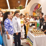 Fotografía A Color Tomada Desde Un Ángulo Bajo, Que Captura Un Recorrido Oficial Por Un Stand De Feria. En El Centro, El Gobernador Javier May Rodríguez, Vestido Con Una Guayabera Blanca Bordada, Sonríe A La Cámara. A Su Lado, Aurora Raleigh De La Cruz Viste Una Camisa Azul Con Flores Blancas.