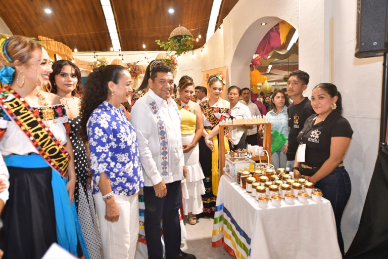 Fotografía A Color Tomada Desde Un Ángulo Bajo, Que Captura Un Recorrido Oficial Por Un Stand De Feria. En El Centro, El Gobernador Javier May Rodríguez, Vestido Con Una Guayabera Blanca Bordada, Sonríe A La Cámara. A Su Lado, Aurora Raleigh De La Cruz Viste Una Camisa Azul Con Flores Blancas.