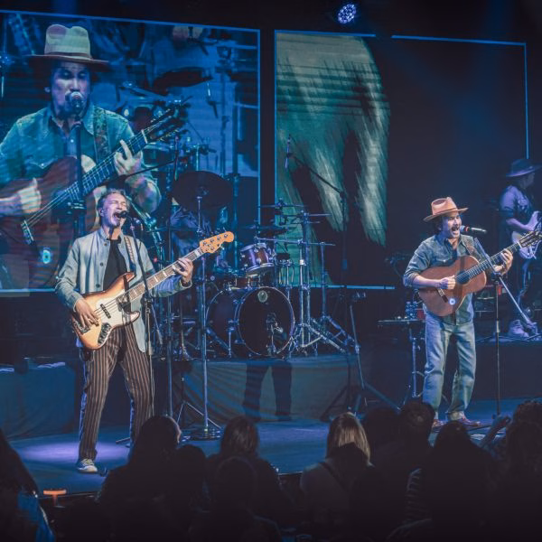 ¡Completamente Gratis!: Bacilos Encabezará El Espectacular Cierre Del Festival Villahermosa 2026 71 Una Toma De Concierto En Vivo Con Un Escenario Oscuro Y Dos Músicos En Primer Plano. El Bajista De La Izquierda Canta Y Toca Frente A Una Pantalla De Video Grande Que Muestra Una Imagen Ampliada De Él. El Guitarrista De La Derecha, Con Un Sombrero Fedora, Toca Y Canta Frente A Otra Pantalla De Video Grande Con Su Propia Imagen.