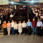 Fotografía En Formato Horizontal Y Plano General Tomada Dentro De Un Auditorio O Planetario Con Gradas Escalonadas. En Primer Plano, Una Línea De Diez Autoridades Y Organizadores (Hombres Y Mujeres Vestidos Con Ropa Formal Y Casual) Posan De Pie Mirando Hacia La Cámara. Detrás De Ellos, Las Butacas Del Recinto Se Encuentran Completamente Llenas Por Cientos De Jóvenes Estudiantes, La Mayoría Vistiendo Uniformes Escolares Blancos Con Pantalones O Faldas Oscuras.