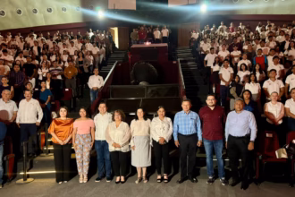 Fotografía En Formato Horizontal Y Plano General Tomada Dentro De Un Auditorio O Planetario Con Gradas Escalonadas. En Primer Plano, Una Línea De Diez Autoridades Y Organizadores (Hombres Y Mujeres Vestidos Con Ropa Formal Y Casual) Posan De Pie Mirando Hacia La Cámara. Detrás De Ellos, Las Butacas Del Recinto Se Encuentran Completamente Llenas Por Cientos De Jóvenes Estudiantes, La Mayoría Vistiendo Uniformes Escolares Blancos Con Pantalones O Faldas Oscuras.