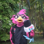 Primer Plano De La Mascota &Quot;10 Lora Comunicadora&Quot; Posando En Los Jardines Del Centro Cultural Quinta Grijalva. La Mascota Es Una Lora Rosa Con Anteojos Negros, Chaleco De Reportera Con El Logo De Canal 10 Y Sostiene Un Micrófono Institucional.