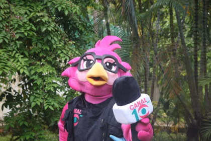 Primer Plano De La Mascota &Quot;10 Lora Comunicadora&Quot; Posando En Los Jardines Del Centro Cultural Quinta Grijalva. La Mascota Es Una Lora Rosa Con Anteojos Negros, Chaleco De Reportera Con El Logo De Canal 10 Y Sostiene Un Micrófono Institucional.