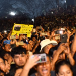 Una Multitud De Personas Disfruta De Un Concierto Nocturno En La Feria Tabasco 2026. Los Asistentes Levantan Sus Teléfonos Celulares Para Grabar El Espectáculo, Creando Puntos De Luz Entre La Audiencia. En El Centro, Destaca Un Fan Que Sostiene Una Cartulina Amarilla Fluorescente Con La Frase &Quot;Tabasco Ama A...&Quot; Y Una Fotografía Del Grupo Musical. El Ambiente Es De Celebración Y Entusiasmo Colectivo.