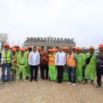 Fotografía De Plano Entero Al Aire Libre Bajo Un Cielo Nublado. En El Centro, La Presidenta Claudia Sheinbaum Pardo, Con Blusa Guinda Bordada, Y El Gobernador Javier May Rodríguez Posan Junto A Una Comitiva De Ingenieros Civiles, Mandos Y Un Numeroso Grupo De Trabajadores De La Construcción Que Visten Uniformes Fosforescentes De Color Verde Y Chalecos Reflejantes Con Cascos Naranjas. Al Fondo Se Observa La Estructura De Concreto En Construcción De Un Puente Elevado De La Carretera Macuspana–Escárcega Y Un Camión De Carga Blanco De La Empresa Supertrack.