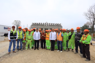 Fotografía De Plano Entero Al Aire Libre Bajo Un Cielo Nublado. En El Centro, La Presidenta Claudia Sheinbaum Pardo, Con Blusa Guinda Bordada, Y El Gobernador Javier May Rodríguez Posan Junto A Una Comitiva De Ingenieros Civiles, Mandos Y Un Numeroso Grupo De Trabajadores De La Construcción Que Visten Uniformes Fosforescentes De Color Verde Y Chalecos Reflejantes Con Cascos Naranjas. Al Fondo Se Observa La Estructura De Concreto En Construcción De Un Puente Elevado De La Carretera Macuspana–Escárcega Y Un Camión De Carga Blanco De La Empresa Supertrack.